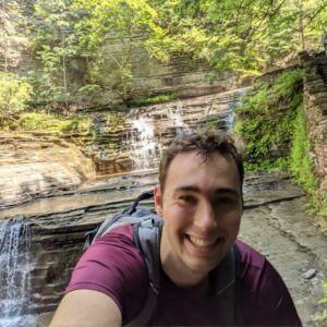 Luke Omodt smiles at camera while wearing a maroon shirt. Little waterfalls and greenery are behind him. 